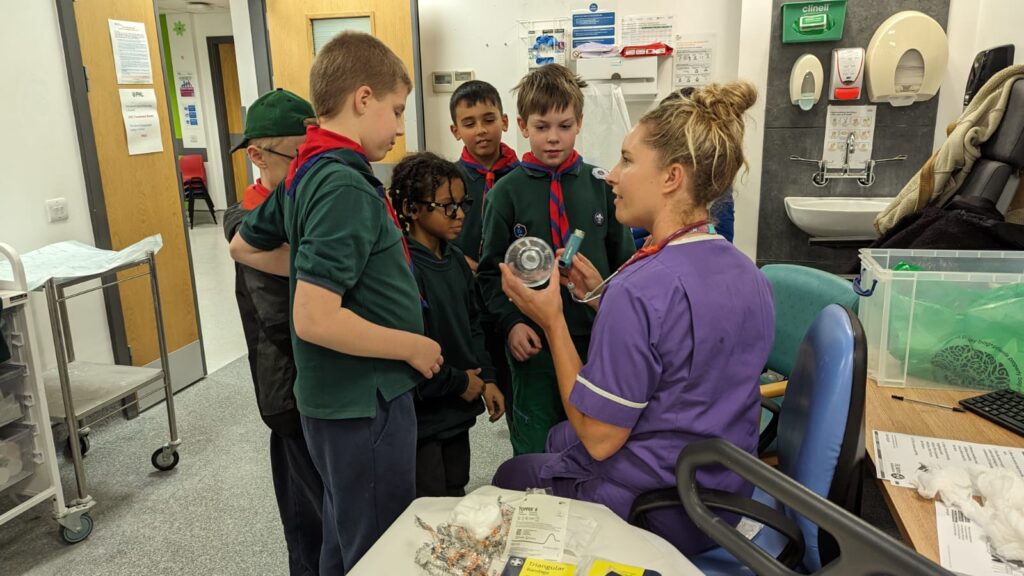 Group of five cubs in a hospital treatment room with a member of hospital staff, who is showing them an inhaler and spacer